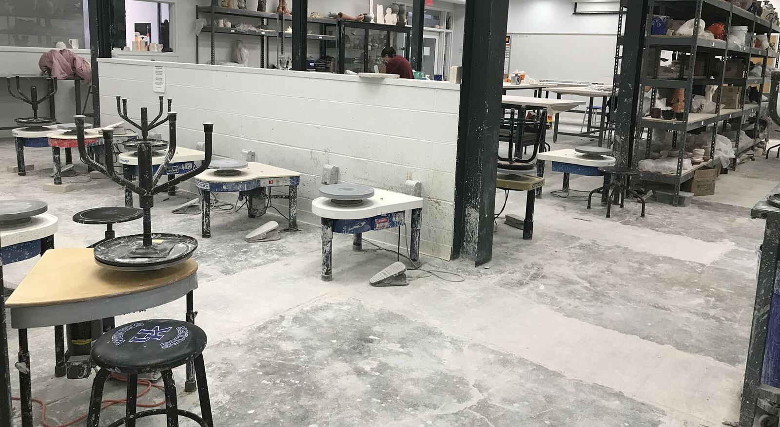 A wide view of the ceramics studio were rows of several throwing wheels are lined up together, shelves are along the back wall for works in progress, and more shelves farther away hold supplies.