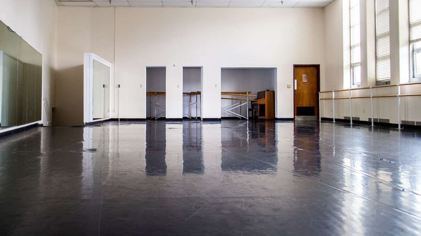 Inside the Charles F. Dickins Dance Studio from the doorway. A mirror wall is on the left, some storage space and a piano in ther back, and a wall of windows with dance bars on the right.