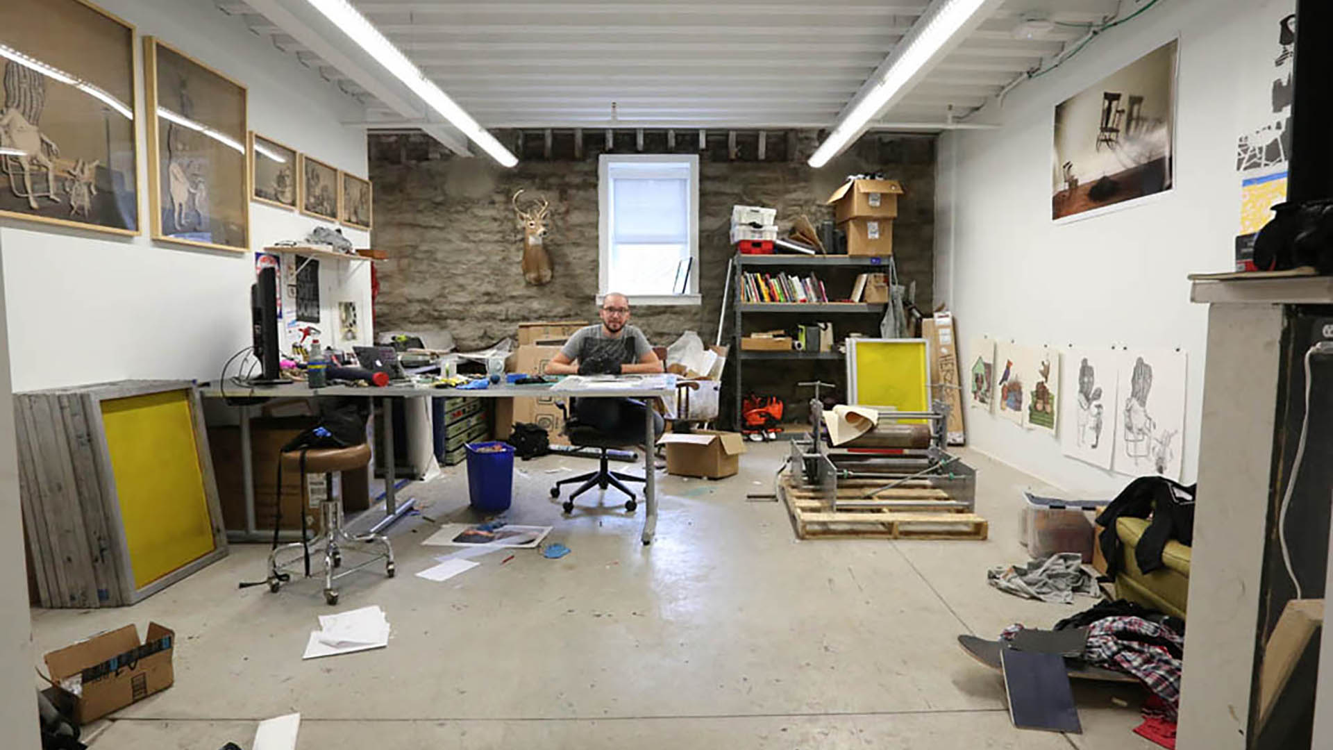 A graduate student sits in their studio with a work in progress in front of them. Various pieces, completed and also in progress, line the walls. Materials and equipment fill shelves and spaces on the floor.
