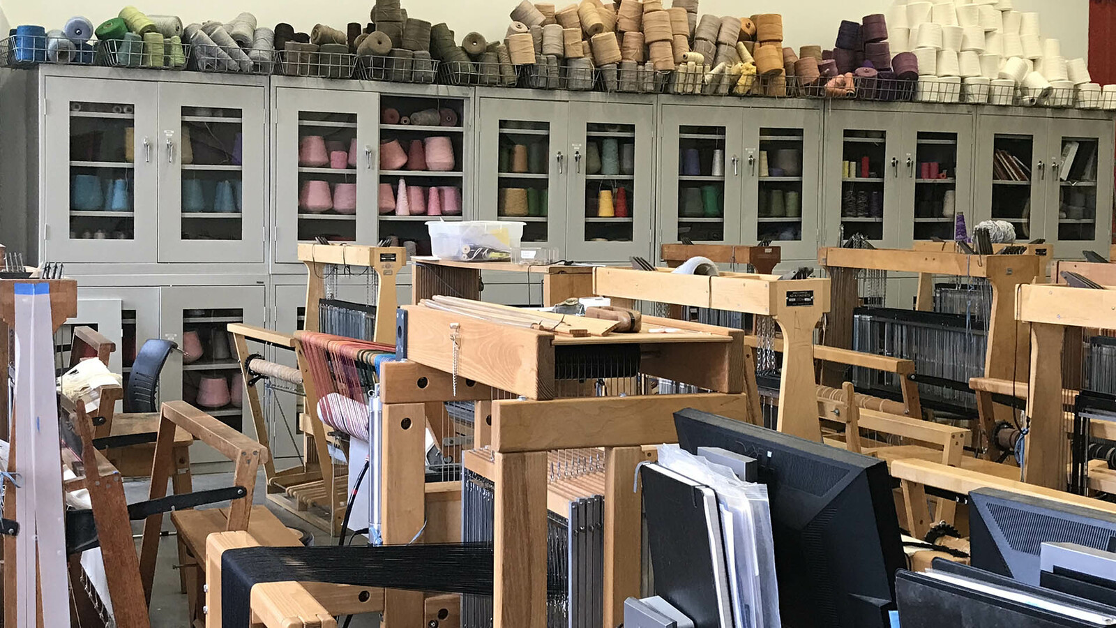 Looking over a room packed with floor looms, many of which have works in progress warped on. Against the back wall are an extensive set of glass door cabinets packed with a rainbow of industrial sized cones of thread and yarn. More colors and sizes of cones are piled on top of cabinets.