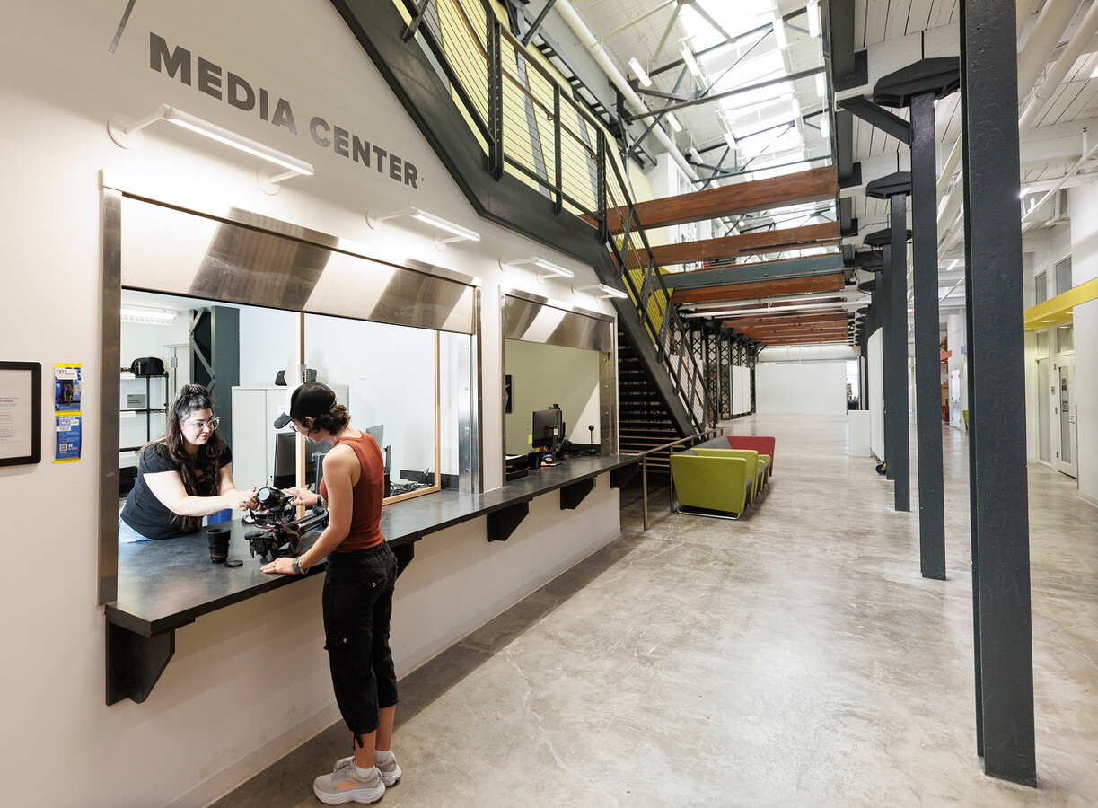 Two open counter bays sit under the main stairs of the Art and Visual studies building with "Media Center" printed above the first counter. A student worker at one counter assists another student in checking out some photography equipment.