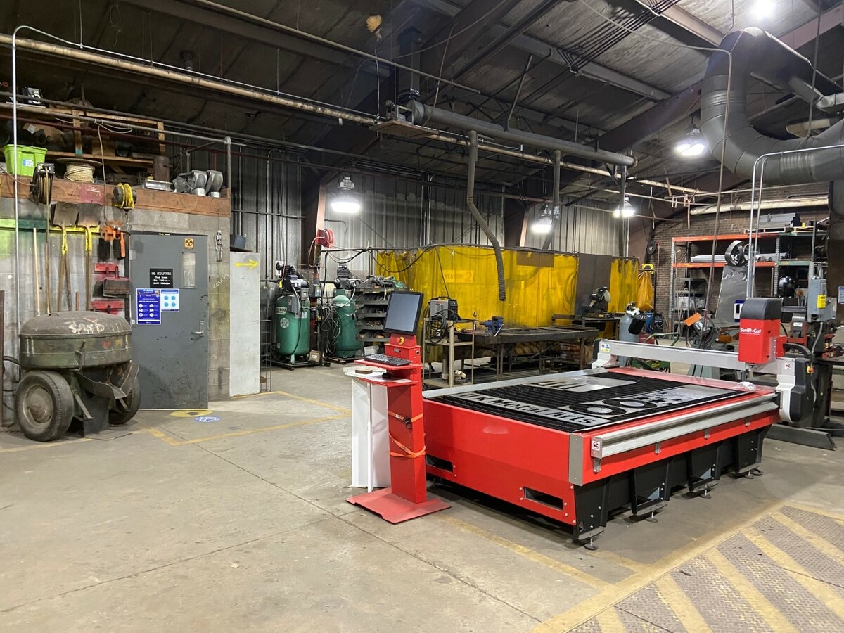 Interior of a large metal building. A bright red digital plasma cutting table is nearest the viewer; with many gas tanks, welders, tables, and other equipment to work with metal in the background.