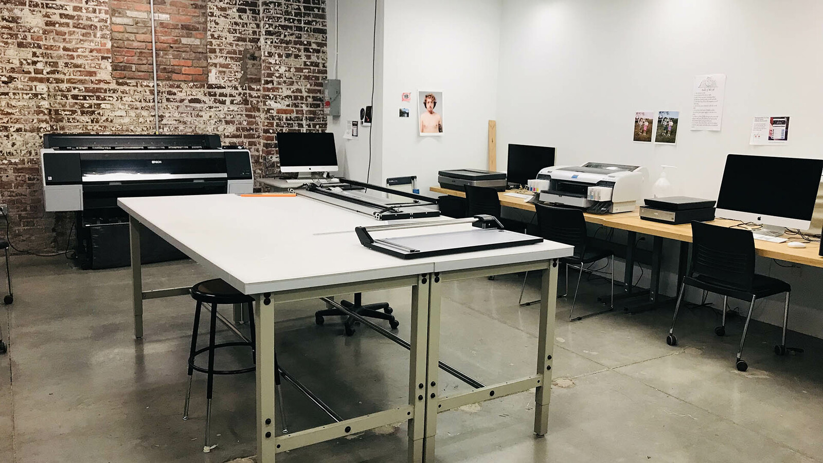 The Digital Printing labl in the photography suite. A large table in the center of the room provides workspace with large paper cutters. Against the right wall are several desks with compters and smaller printers. The back brick wall is taken up by a large format printer.