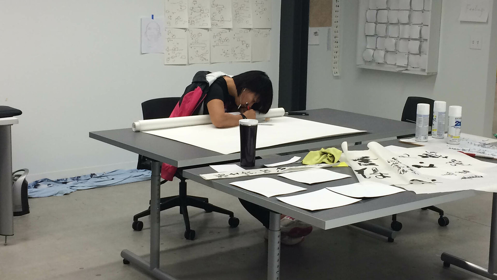 A studnet works on a large rolled out sheet of paper at a table in the middle of a white room. Taped up on the wall behind them are several sketches and iterations on designs.