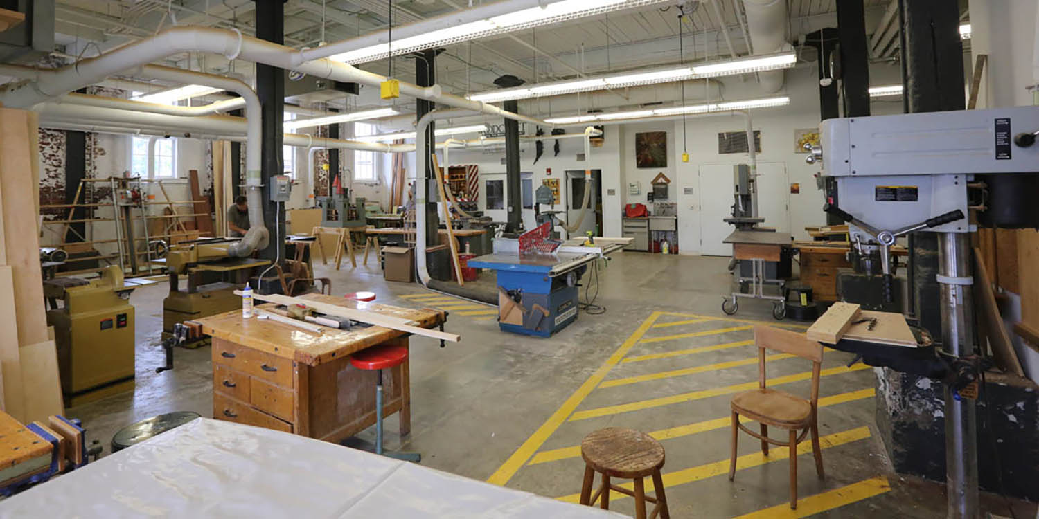 A huge warehouse-like room with copious ventilation ducts and lightning. A drill press is on the close right, and in the distance a tablesaw, planar, bansaw, jointer, and other heavy equipment can be seen. Yellow painted caution zones are marked generously around each piece of equipment.