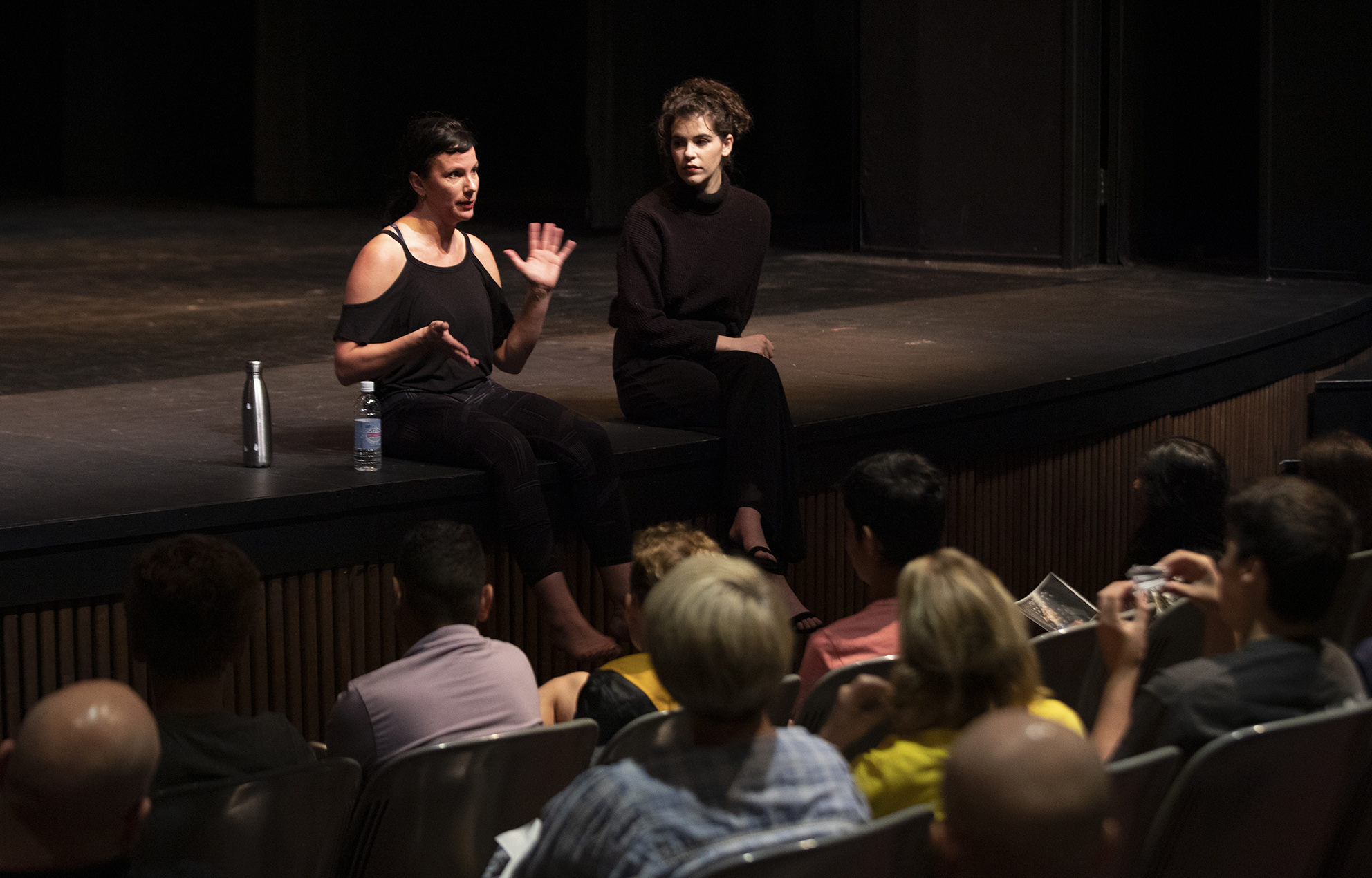two people sitting on stage talking to students in theater seats