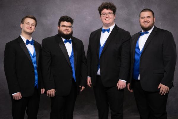 Members of FreshF stand against a grey background wearing tuxedos with blue bowties and vests 
