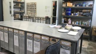 A large high metal table takes up the center of the room with vertical drawers below labled for various glaze and pigment materials. A tile color swatch board is on the far back wall and several shelves with more supplies.