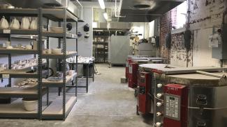 Several electric kilns line the right wall, with many shelves of works in progress lining the left wall. More shelves and storage continue on the far wall.