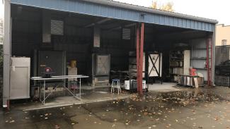A large open air outdoor metal shed with blue and red paint. Set against the back wall are several large gas kilns with a few metal folding tables scattered around the space for loading and unloading firings.