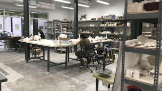 A brightly lit room lined with shelves for projects and supplies. Several large tables are arranged in the center of the room as communal workspace with many stools available.