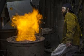 A student stands back from a burning furnace. A pillar of red and gold flame blooms out of the top of the furnace.