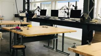 Standing desks with computers, small lightning kits, and other gear line a wall of windows. The room beyond the computers holds several large tables with clamps and spare wiring for extra working space.