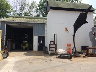 Exterior of the Metal Arts Building, a white and green metal and cinderblock structure. Several in progress sculptures stand to the side and equipment is scattered around them.