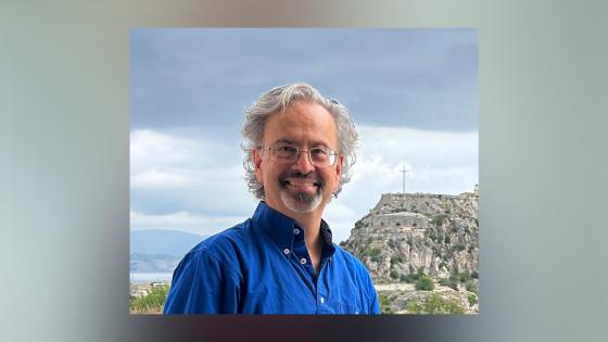 Headshot of Dr. Ben Arnold, wearing a blue button down and glasses against a blue sky 